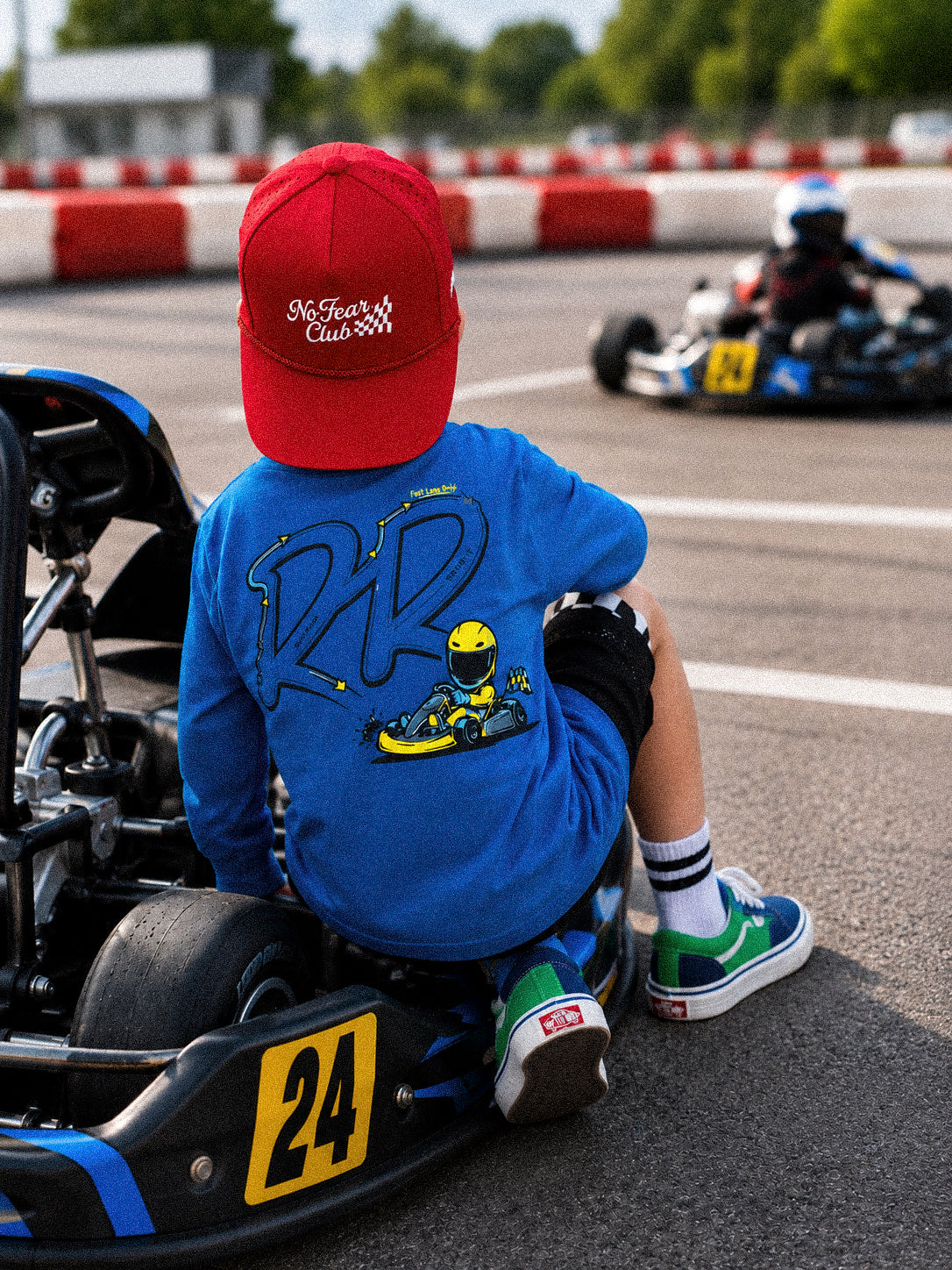 A child wearing a red cap and a blue t-shirt with a go-kart graphic sits on the back of a race number 24 go-kart on a track, with another kart visible in the background.