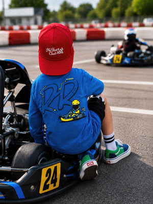 A child wearing a red cap and a blue t-shirt with a go-kart graphic sits on the back of a race number 24 go-kart on a track, with another kart visible in the background.
