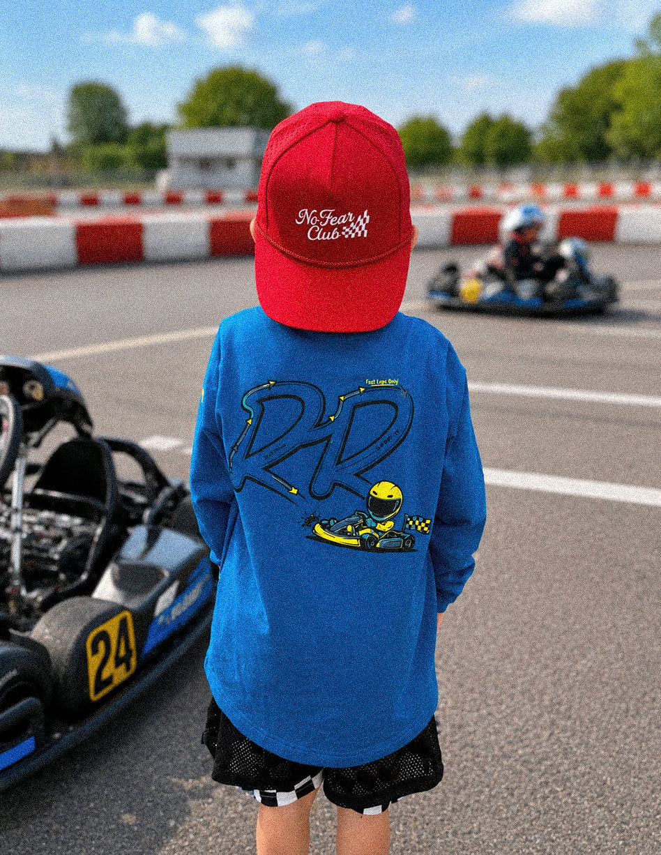 A child standing on a race track wearing a bright blue long-sleeve shirt with a large '22' and karting graphic on the back, a red baseball cap with 'McLaren Cars' text, black shorts, and colorful sneakers. Gokarts are visible in the background.